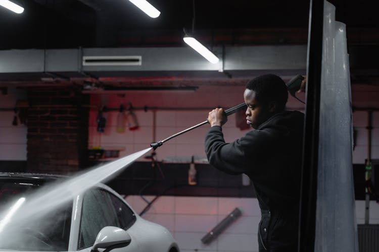 A Man In Black Jacket Spraying Water On A Car