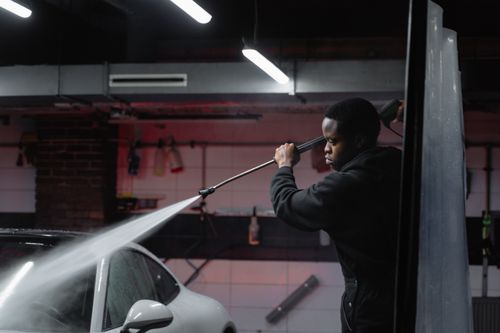 Free A man uses a high-pressure hose to wash a car in an indoor garage setting. Stock Photo