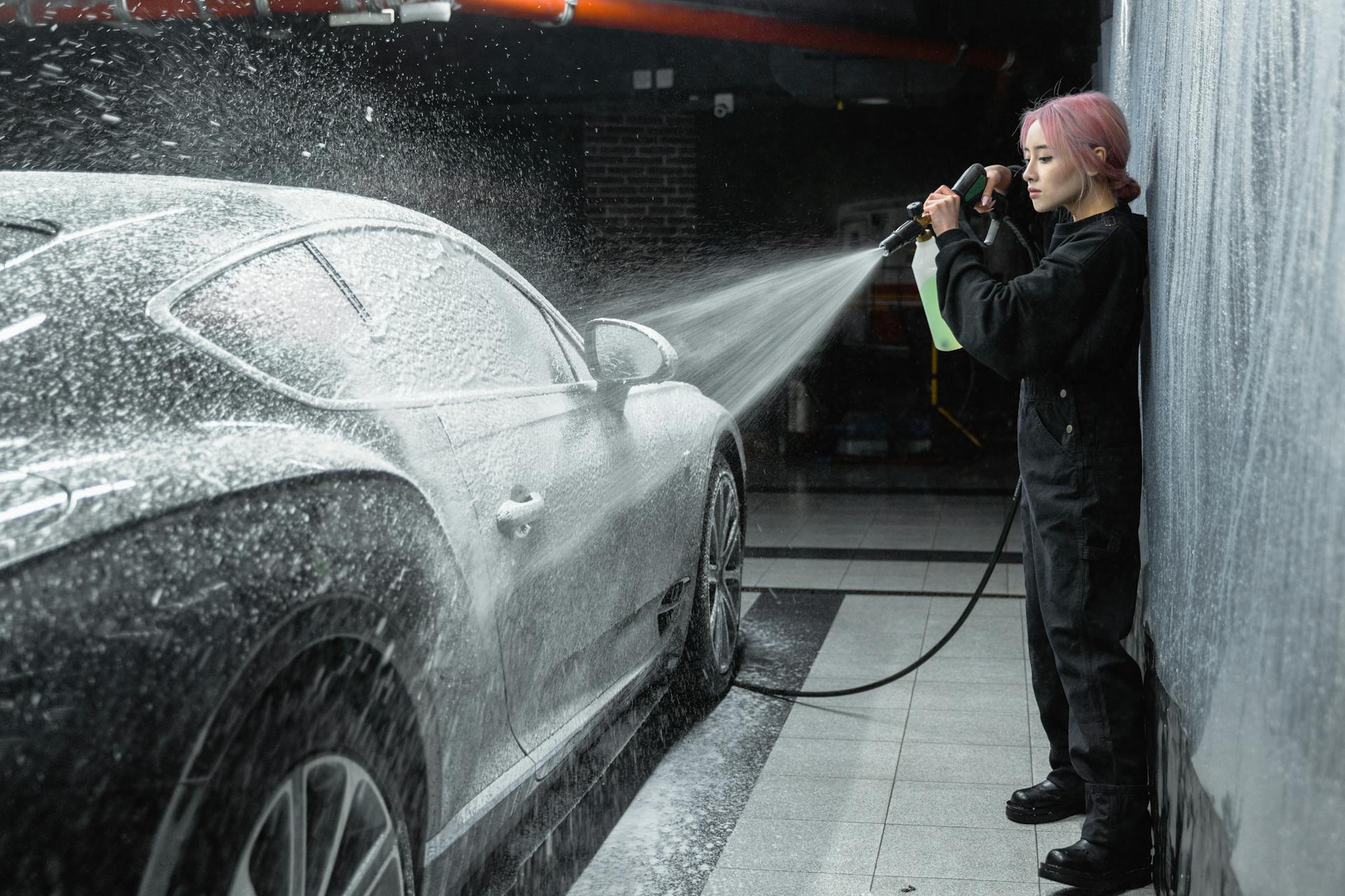 An Asian woman using a pressure washer to clean a car at an indoor car wash facility.