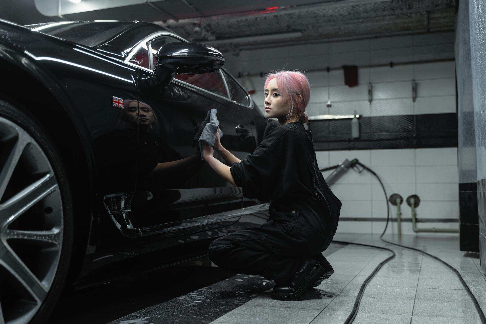 A young female worker polishing a black luxury car in an indoor garage setting.