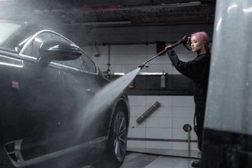 Free Woman power washing a black car in an indoor garage using a high-pressure washer. Stock Photo