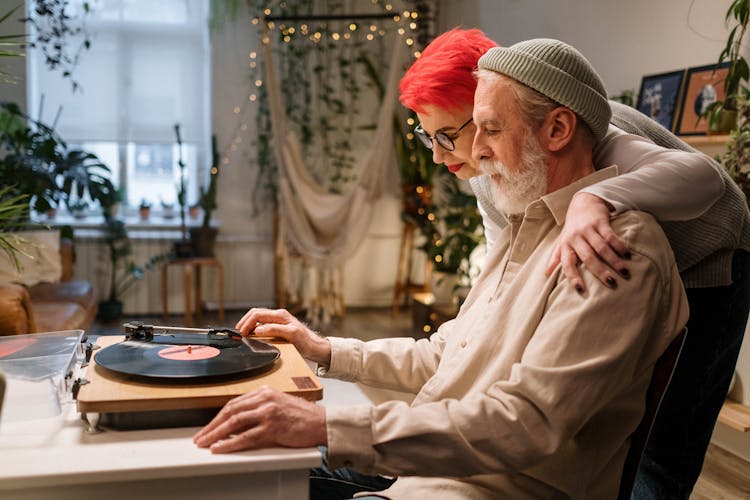 Woman Standing With Arm Around Man Listening To Record Player