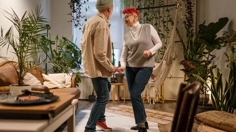 Senior couple dances joyfully in a bohemian home interior filled with indoor plants.