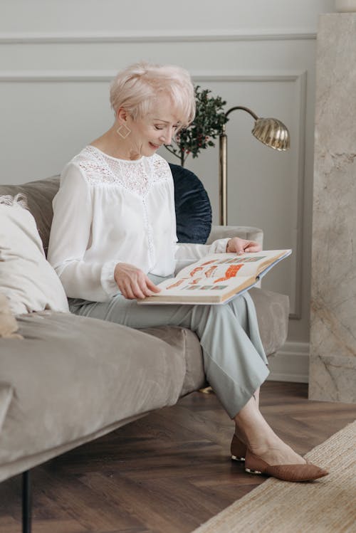 A Woman in White Top Sitting on the Sofa while Reading a Book