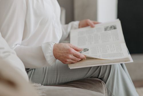 Free A senior adult in white attire reading a book while sitting indoors. Stock Photo