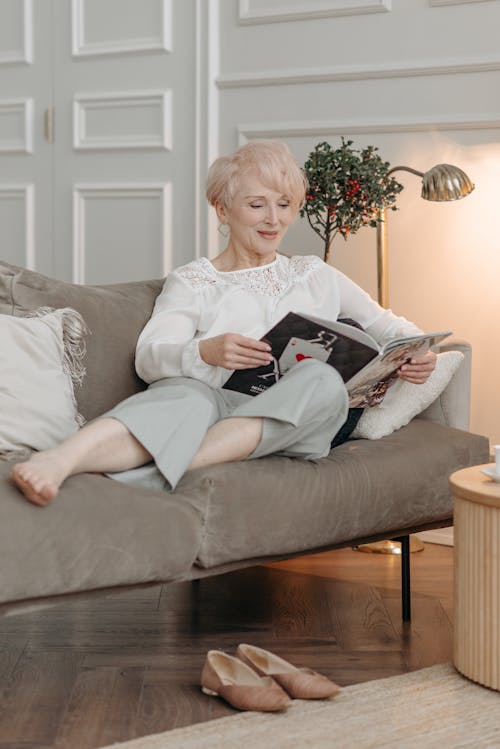 A Woman in White Top Sitting on the Sofa while Reading a Book