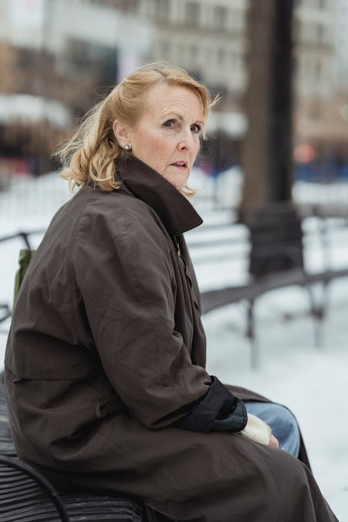 Free Side view of scared senior female in coat looking away while sitting on bench in city Stock Photo