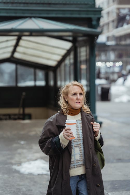 Senior female in coat with disposable cup of hot beverage looking away on city street in winter