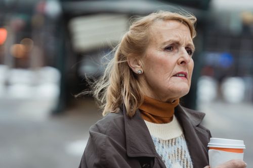 Free Pensive elderly female with takeaway hot drink looking away in town on windy day on blurred background Stock Photo
