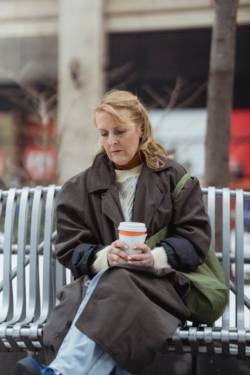 Free Lonely elderly female in coat with hot drink to go sitting with crossed legs on bench while looking down Stock Photo