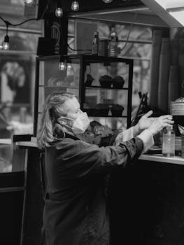 Black and white side view of elderly female customer in protective mask applying antibacterial gel at counter in bakery