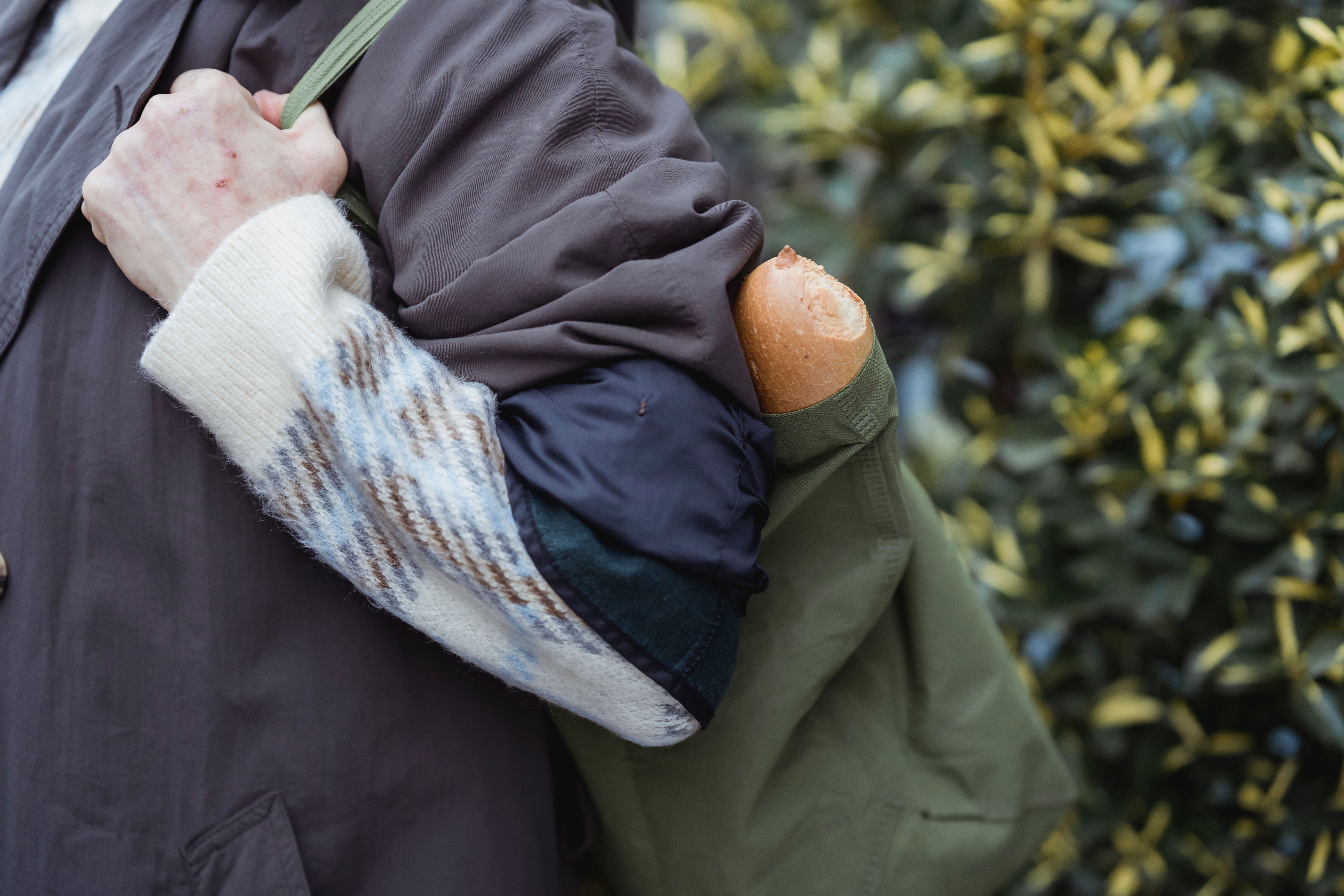 Crop unrecognizable female in sweater and coat with wheat bread in textile bag against plant