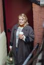 Elderly female in coat with hot drink to go and baguette in bag looking forward against house door in town