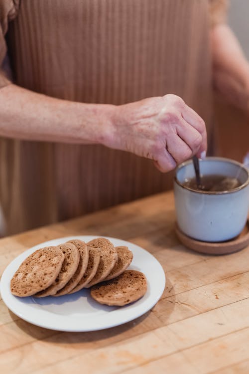 Free Crop unrecognizable female stirring hot drink in mug at table with delicious biscuits in house Stock Photo