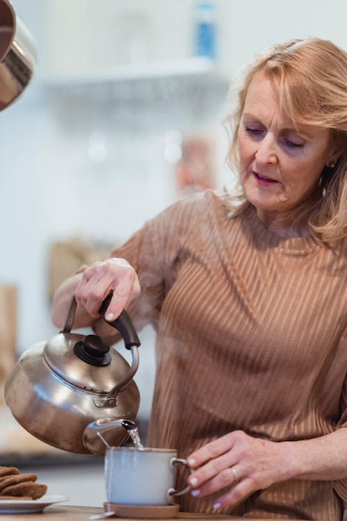 Free Senior female with kettle pouring hot water into mug at table with cookies at home Stock Photo