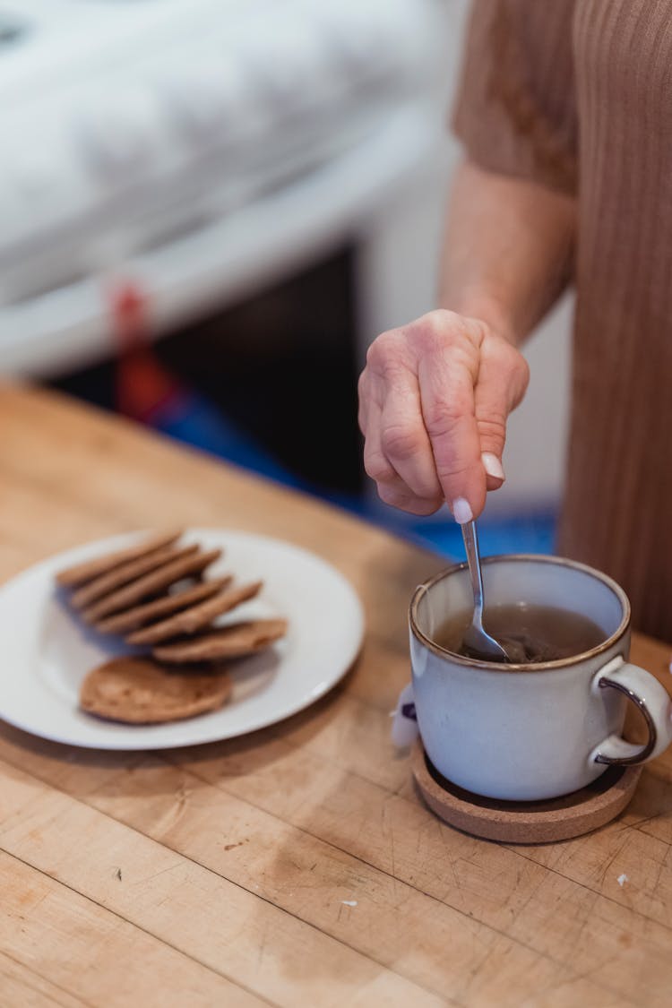 Crop Woman Brewing Tea At Desk With Biscuits In Kitchen