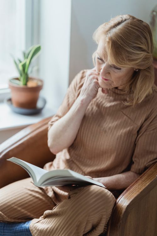 Free Senior female touching face while reading textbook in armchair against window in house room in daytime Stock Photo