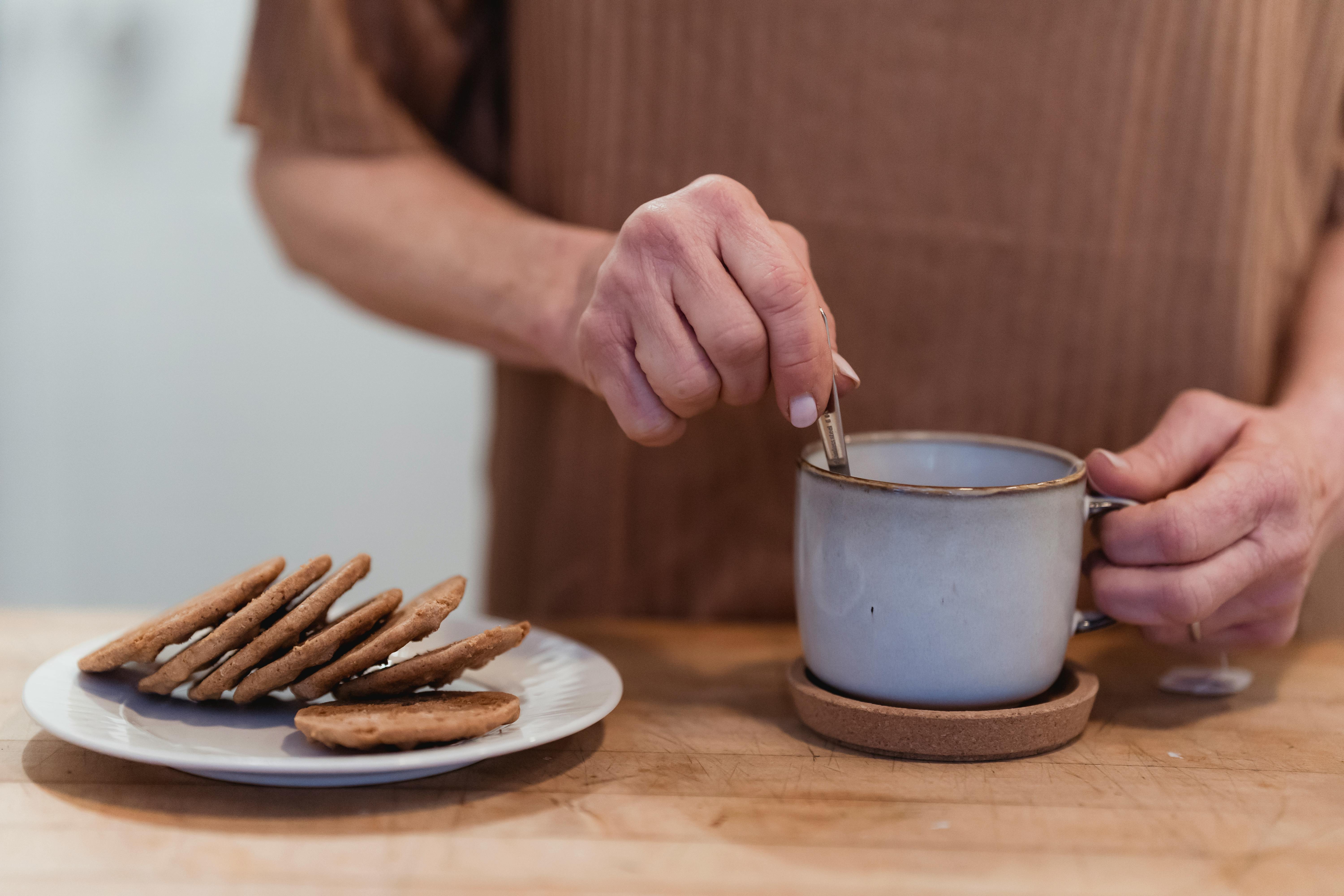 Crop anonymous female preparing hot beverage in mug at desk with tasty biscuits on plate at home
