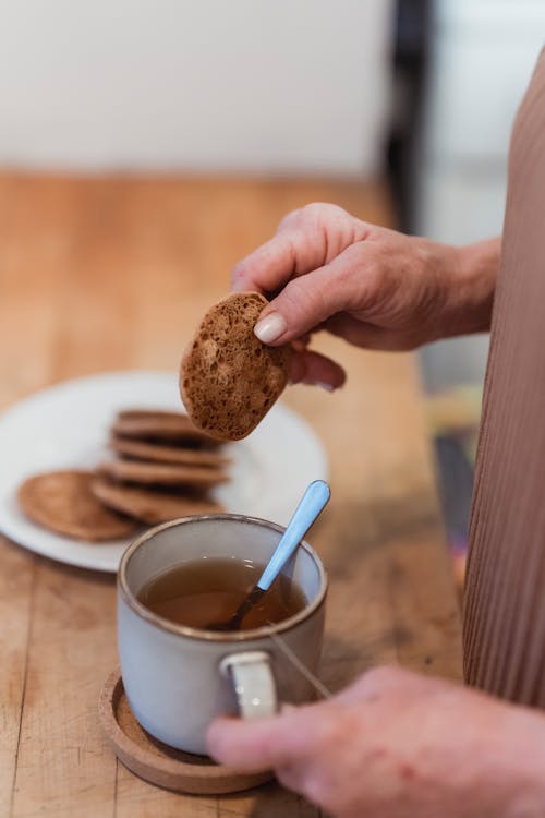 Free Crop unrecognizable female with mug of hot drink and tasty biscuit at table in house Stock Photo