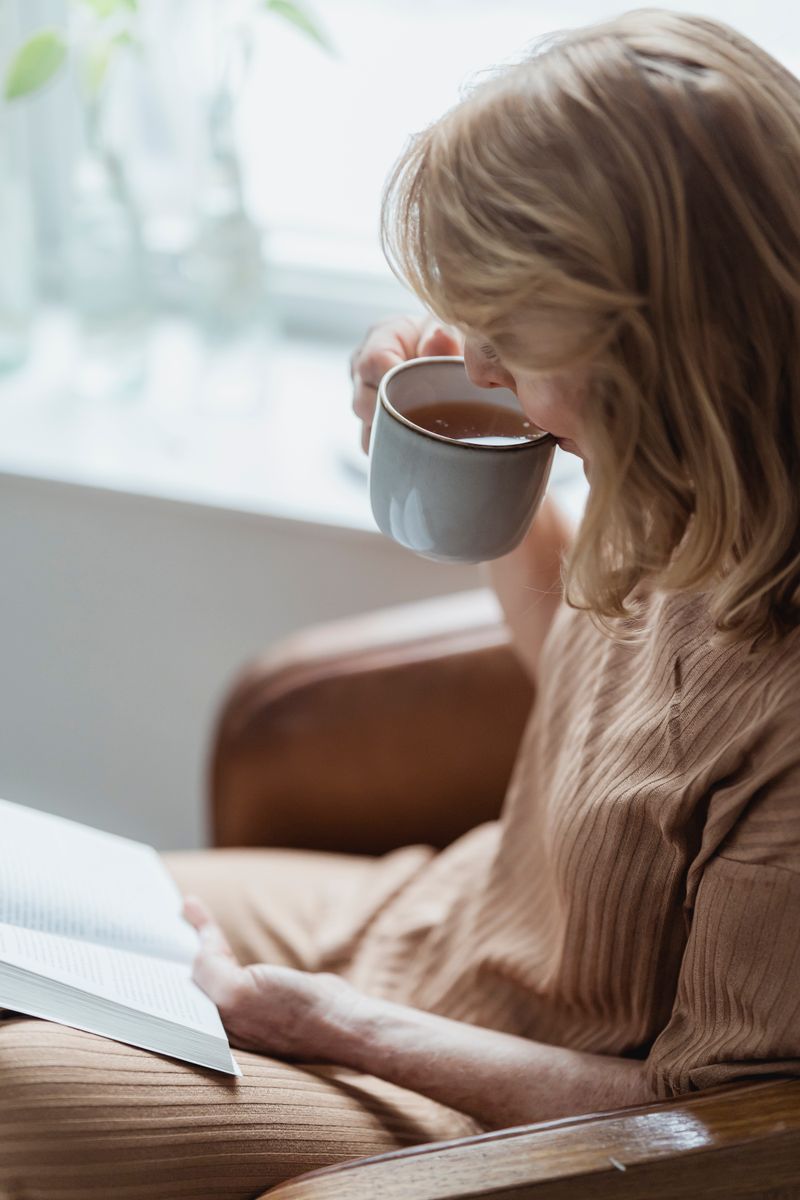 A senior woman resting in an armchair by a sunny window with a book and a cup of tea