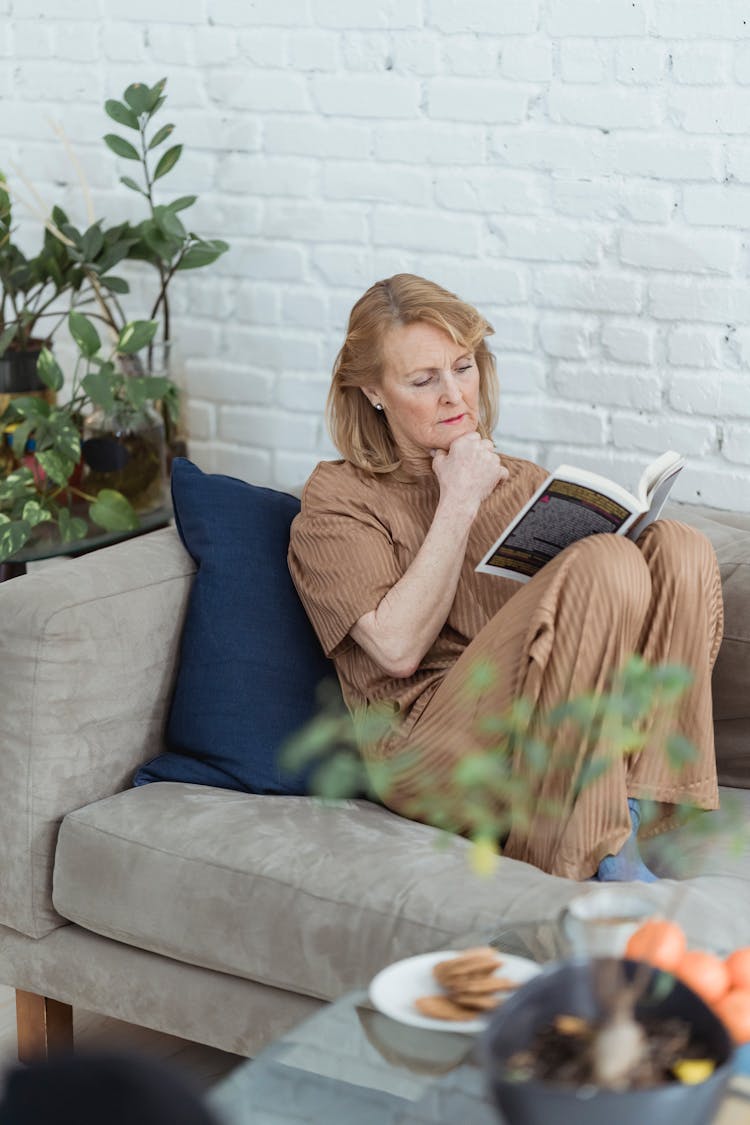 Elderly Woman Reading Book On Sofa At Home