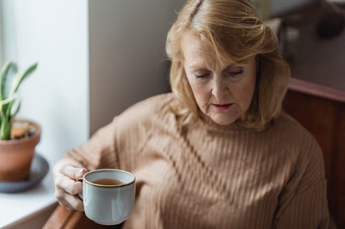 Free Senior concentrated woman sitting near windowsill and drinking tea Stock Photo