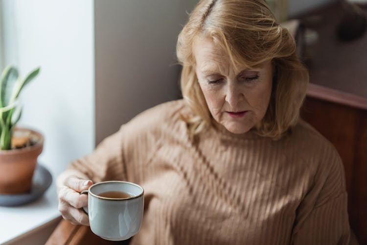 Senior Concentrated Woman Sitting Near Windowsill And Drinking Tea