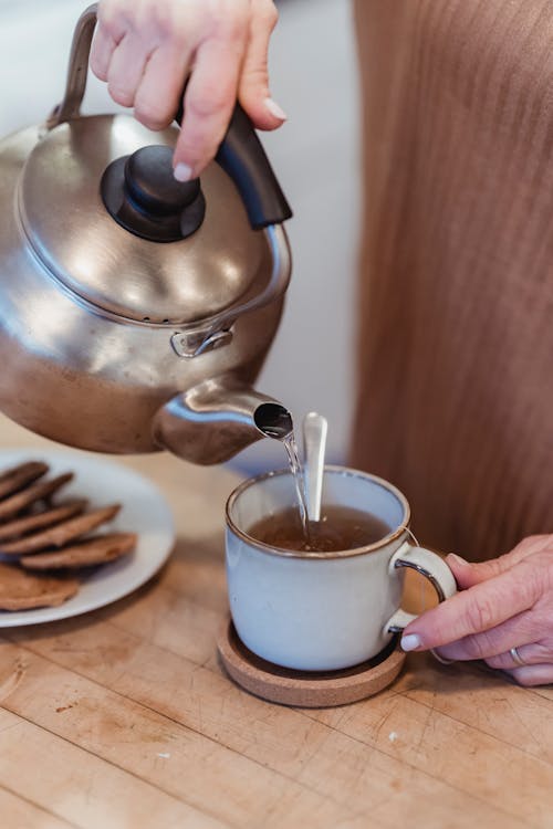 From above of crop anonymous woman pouring hot water from metal kettle into cup while brewing tea for breakfast