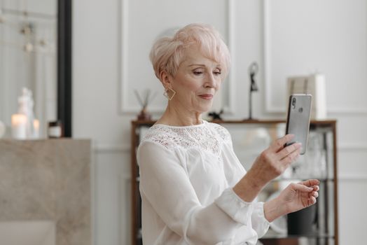 Elegant senior woman in white blouse using a smartphone indoors, embodying technology and elegance.