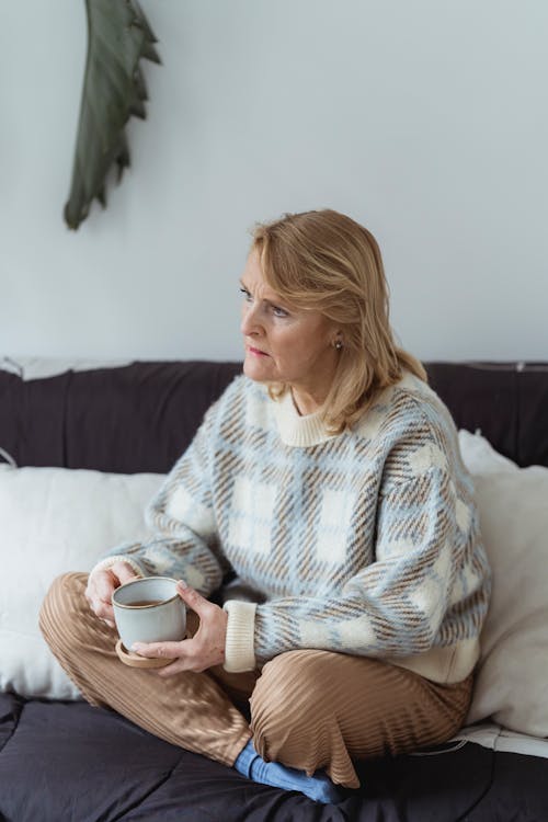 Free Full body of pensive mature female with blond hair in warm sweater drinking cup of coffee while sitting on couch with crossed legs and looking away at home Stock Photo