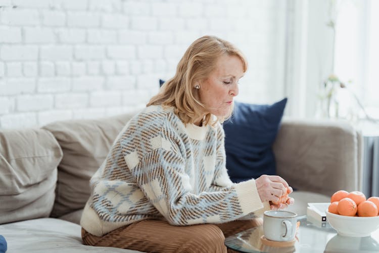 Mature Woman Drinking Coffee And Peeling Mandarins While Sitting On Sofa