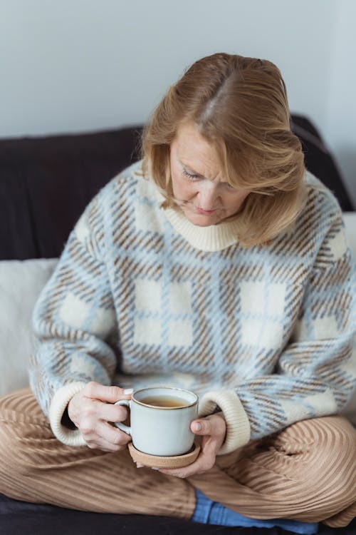 Free Melancholic elderly female with blond hair in warm sweater sitting on comfortable sofa with crossed legs and drinking cup of hot tea during weekend at home Stock Photo