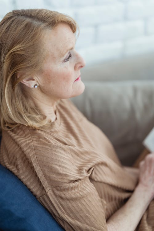 Side view of tranquil mature woman with blond hair sitting on comfy sofa with opened book on knees and looking away dreamily