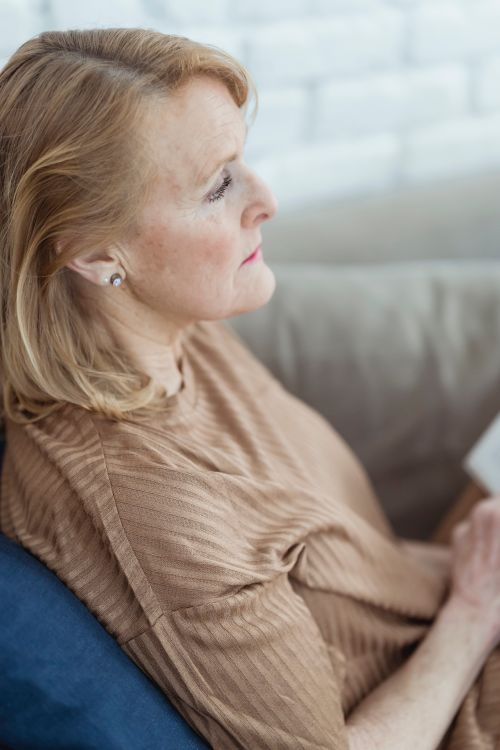 Free Side view of tranquil mature woman with blond hair sitting on comfy sofa with opened book on knees and looking away dreamily Stock Photo