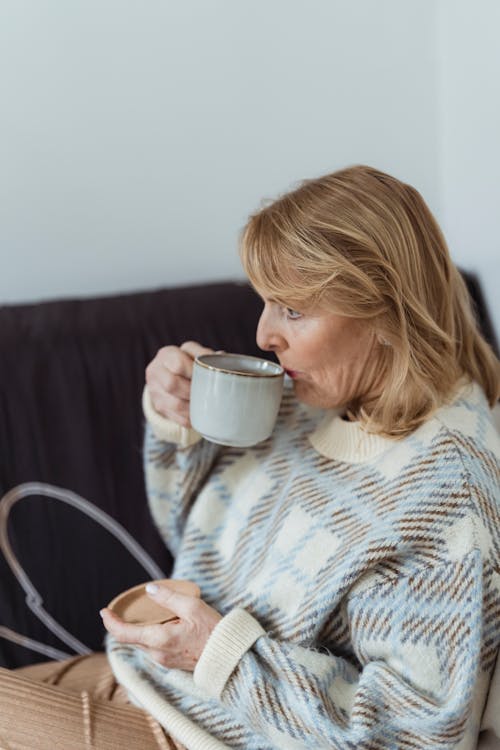Free Side view of relaxed middle aged female with blond hair in warm sweater sitting on sofa and drinking hot aromatic coffee Stock Photo