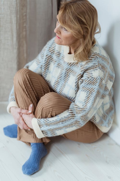 From above of upset middle aged woman in warm sweater embracing knees and looking away thoughtfully while sitting on floor at home