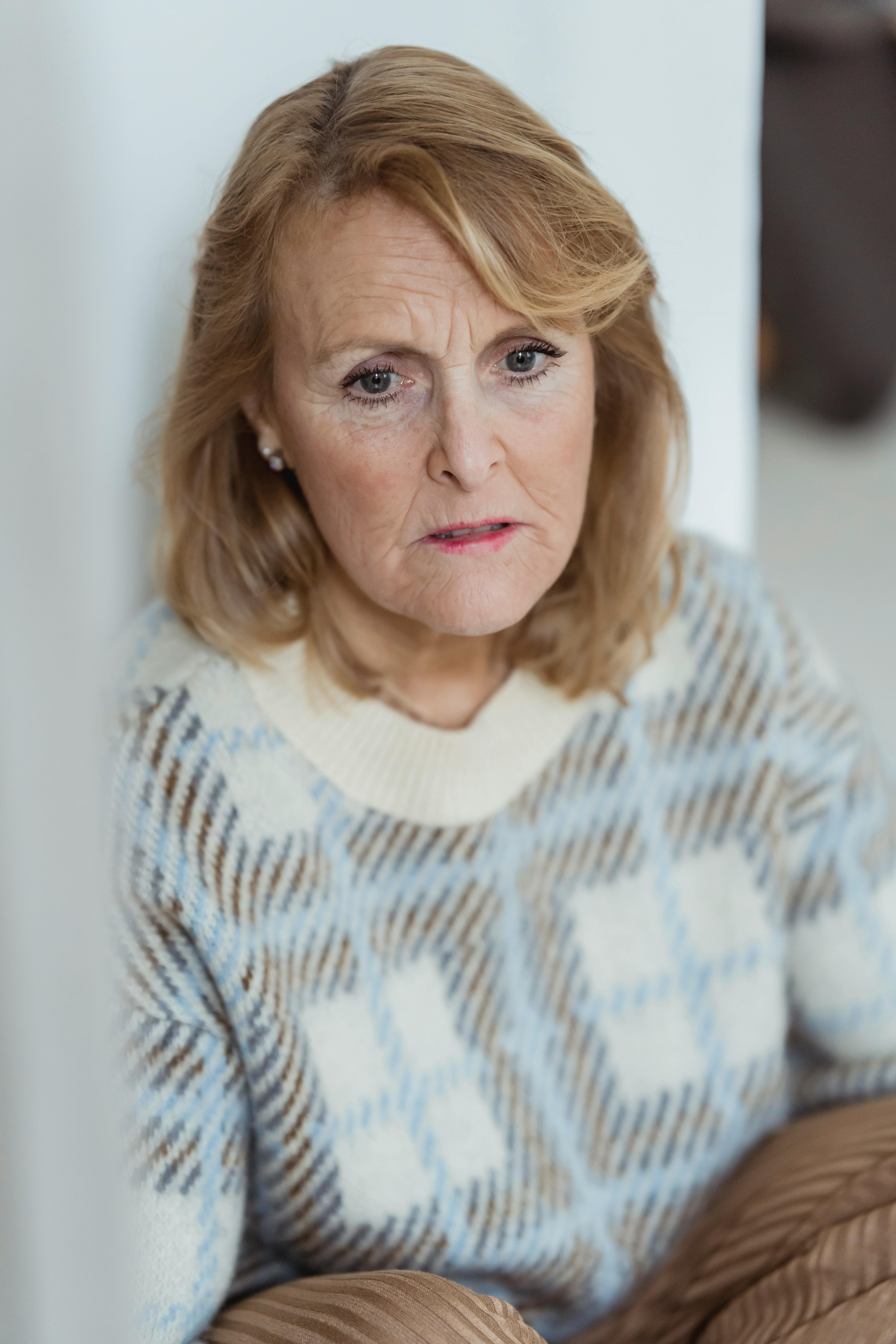 Unhappy mature lady with blond hair in war sweater looking away pensively while sitting on floor near white wall