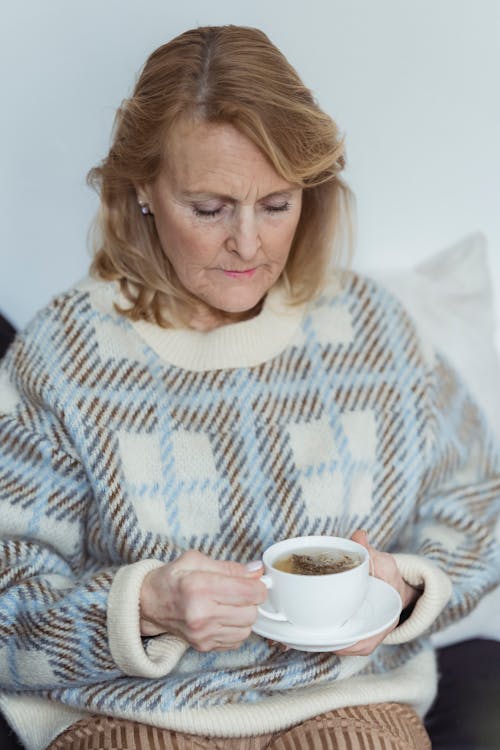 Tranquil mature woman in warm casual outfit drinking cup of freshly brewed tea while resting on couch at home