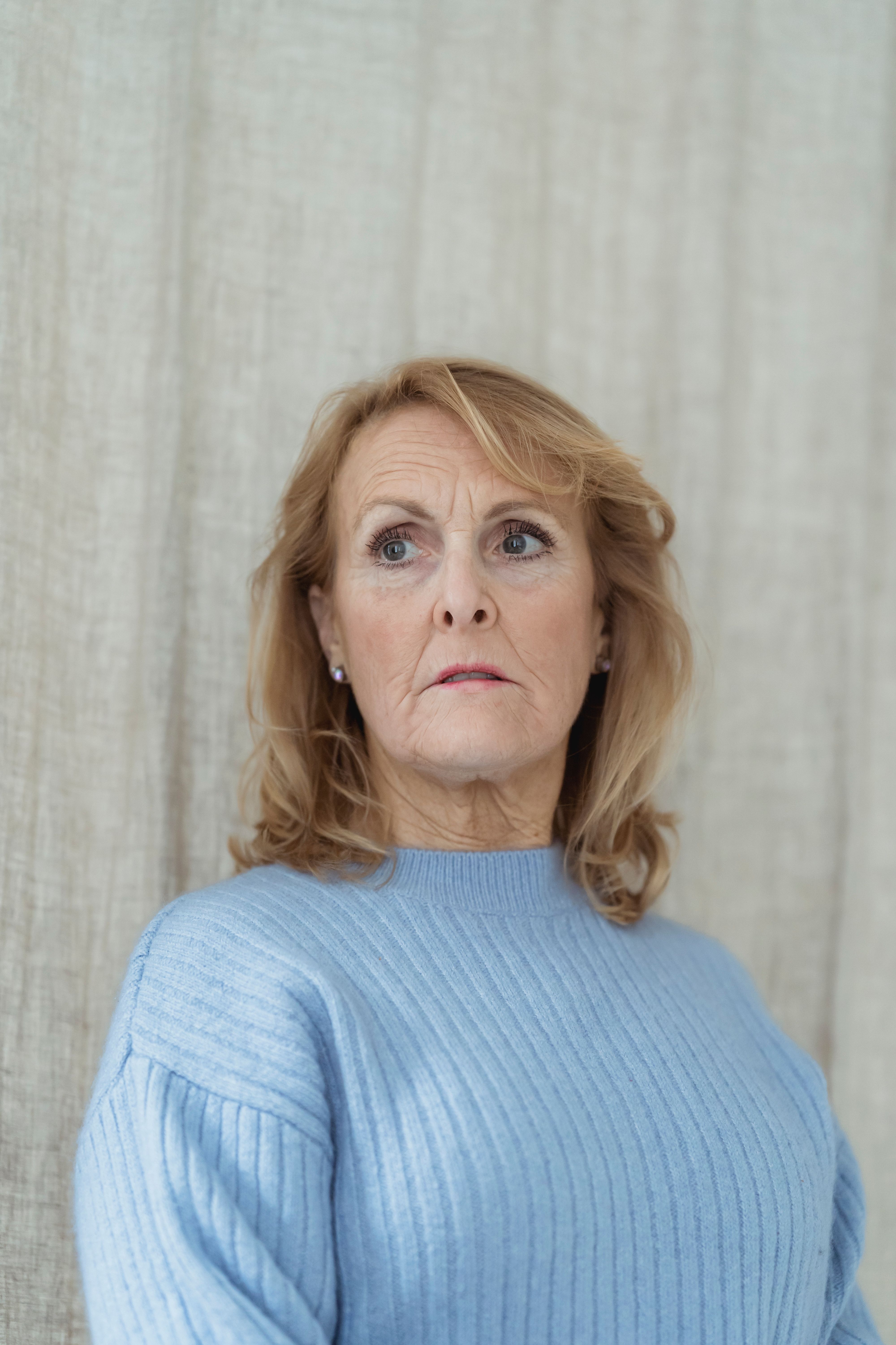 Emotionless middle aged female with wavy blond hair in blue warm sweater standing near curtain and looking away on sunny day at home