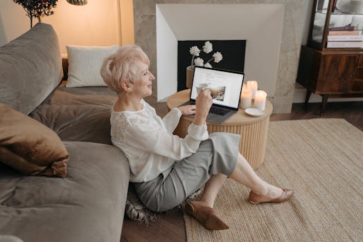 Elderly woman enjoying tea while working remotely on a laptop at home.