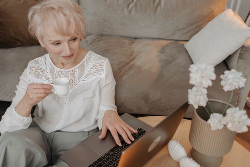 Free Senior woman enjoying coffee and working remotely from home with a laptop, embracing digital life. Stock Photo