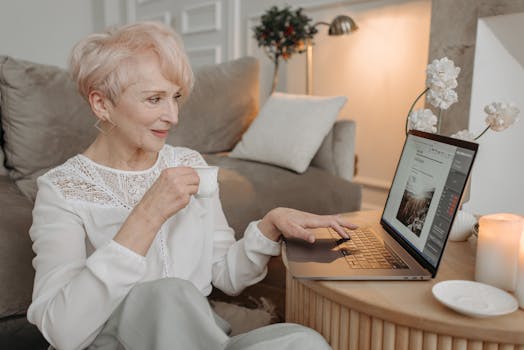 Elderly woman with short hair working from home on a laptop, enjoying coffee.