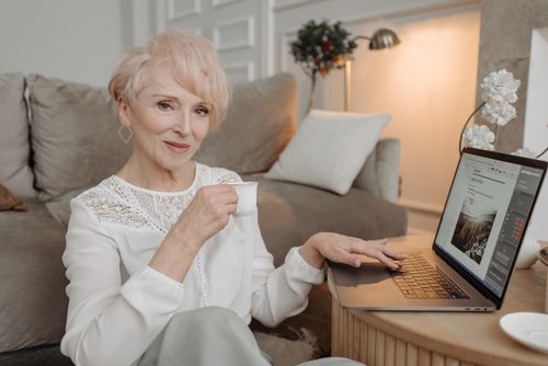 Free Senior woman drinking tea and working on laptop from home, epitomizing comfort in remote work. Stock Photo