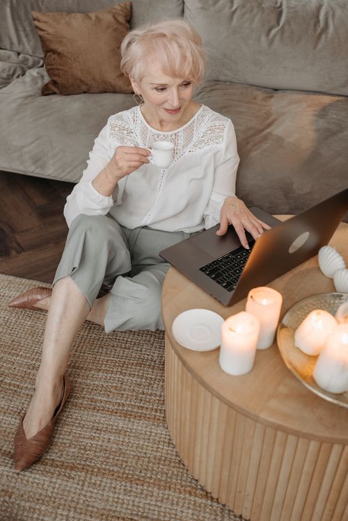 Free Senior woman enjoying remote work from home, sipping tea, using a laptop in a cozy setting with candles. Stock Photo