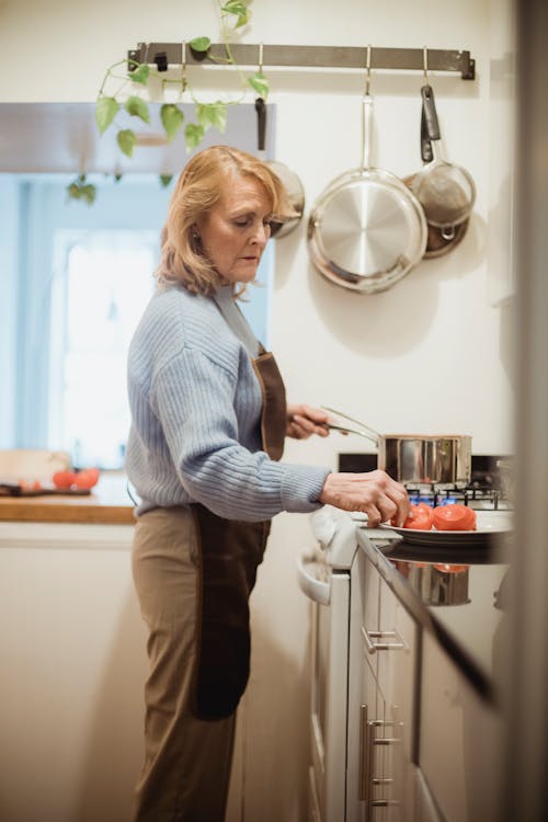 Side view unemotional mature female in apron cooking meal and standing near stove with saucepan in light kitchen