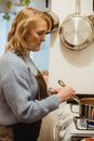 Side view focused senior housewife in blue sweater and apron stirring meal boiling in saucepan placed on stove in light kitchen