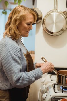 Side view focused senior housewife in blue sweater and apron stirring meal boiling in saucepan placed on stove in light kitchen