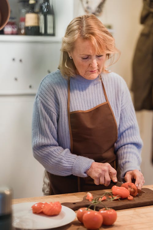 Focused mature female chef in apron cutting fresh ripe tomatoes on chopping board while cooking in kitchen