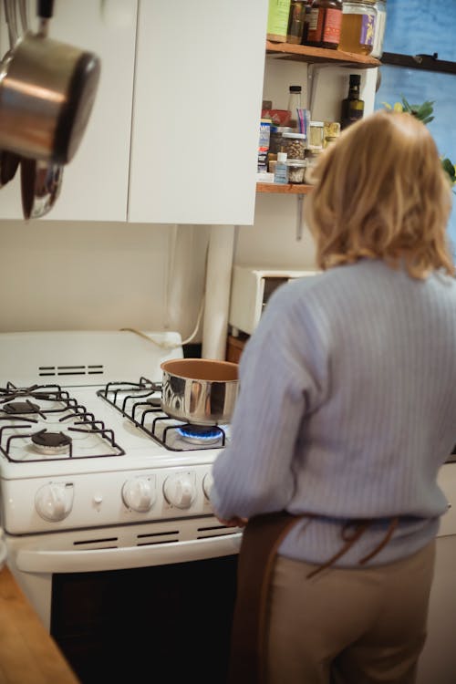 Back view unrecognizable female in apron standing near saucepan placed on stove in kitchen