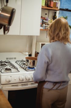 Back view unrecognizable female in apron standing near saucepan placed on stove in kitchen